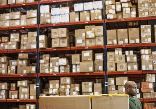 African American warehouse worker checking inventory next to large racks of cardboard boxes holding product in a distribution warehouse.
